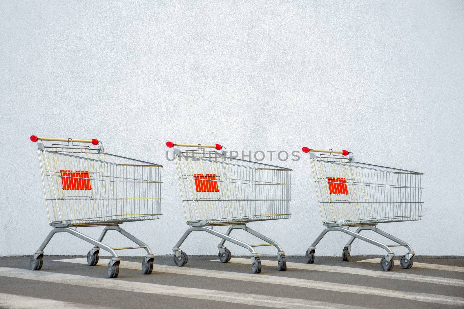 Three Empty Shopping Cart Trolley Stands near Mall with Copy Space. Grocery Cart on the White Wall Store. Trolley at the Supermarket. E-commerce. Shopping Concept. Side View. Black Friday Sale. Online by synel