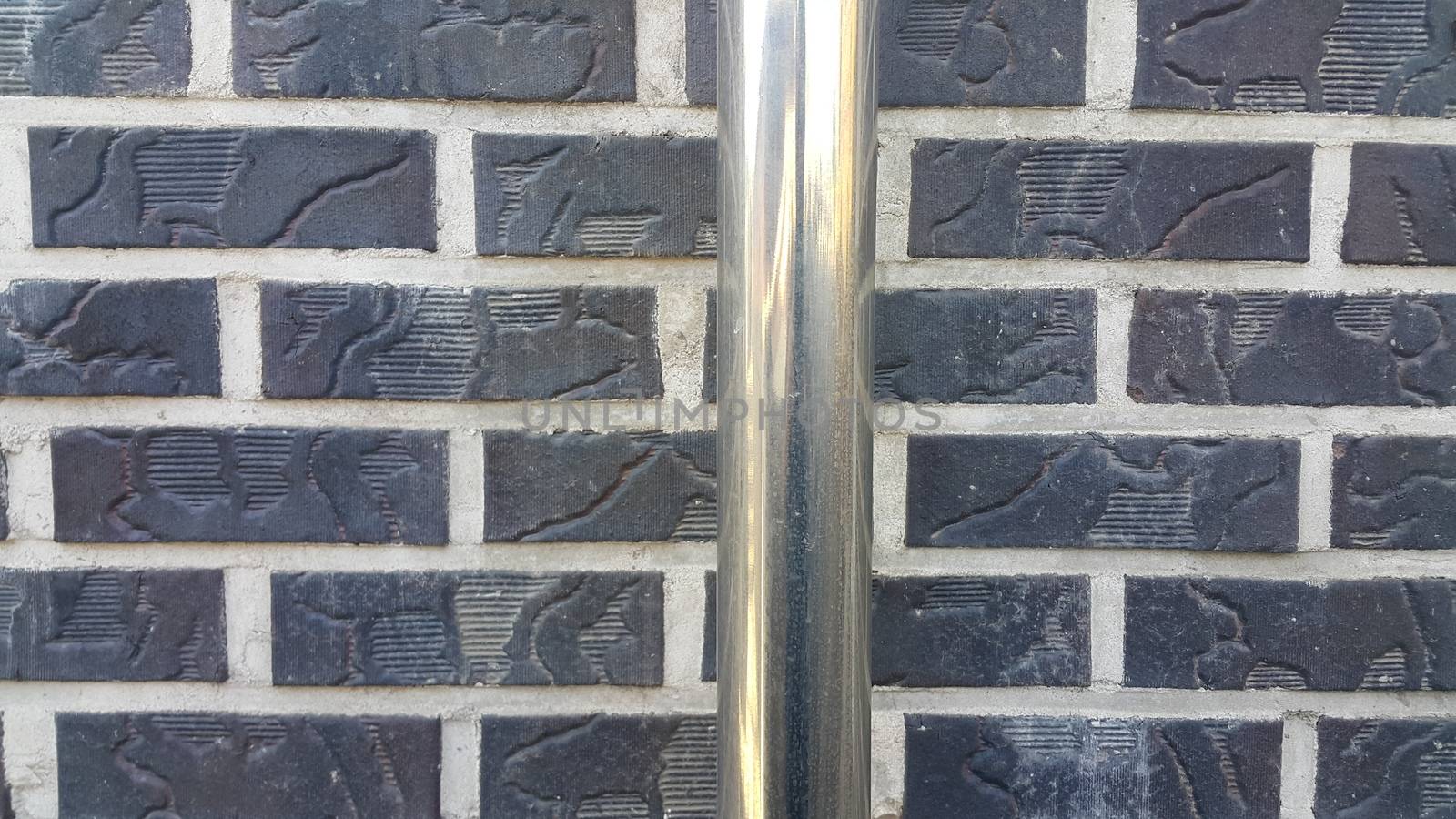 Closeup view of silver water pipe over black brick wall. A background of silver pipe over a weathered old exterior brick wall in the sunshine.