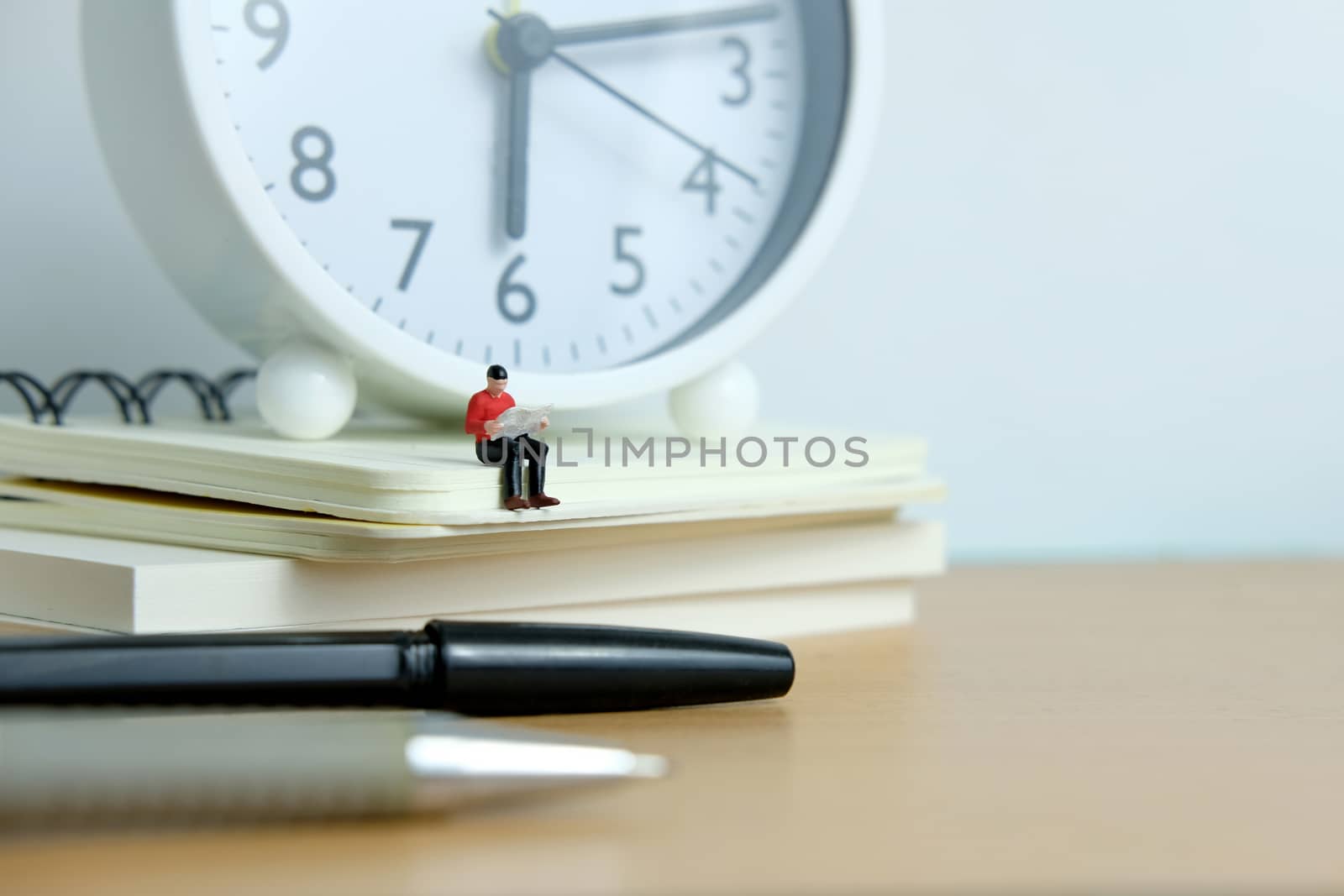 Young man sitting on book pile while reading with pen and white clock