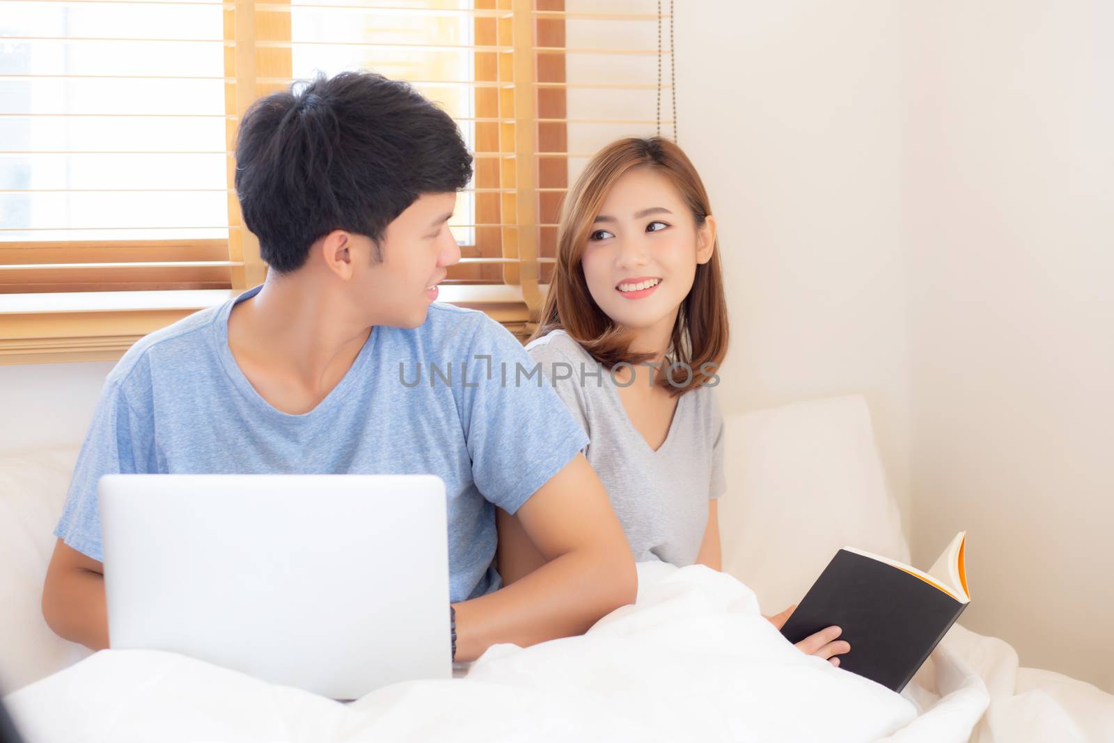 Asian young couple with man using laptop computer and searching internet and woman reading notebook on bed at bedroom with happy and relax, communication and business concept.