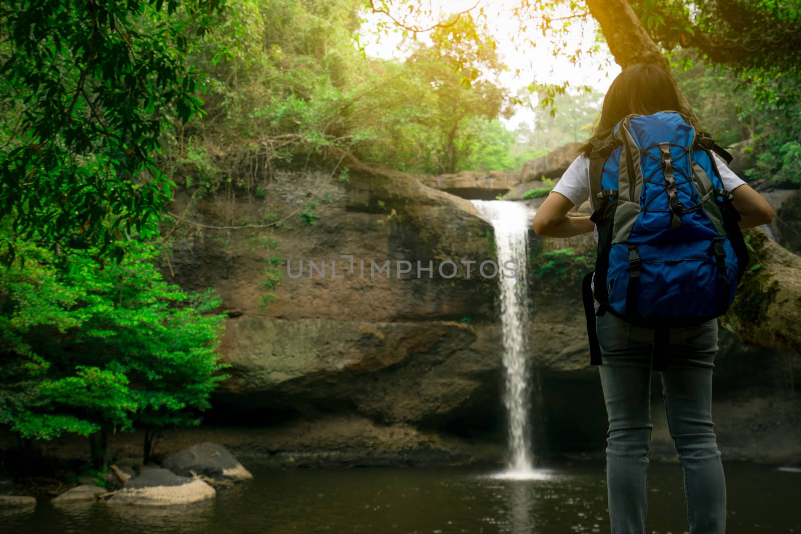 Back view of Asian woman with backpack watching small waterfall  by Fahroni