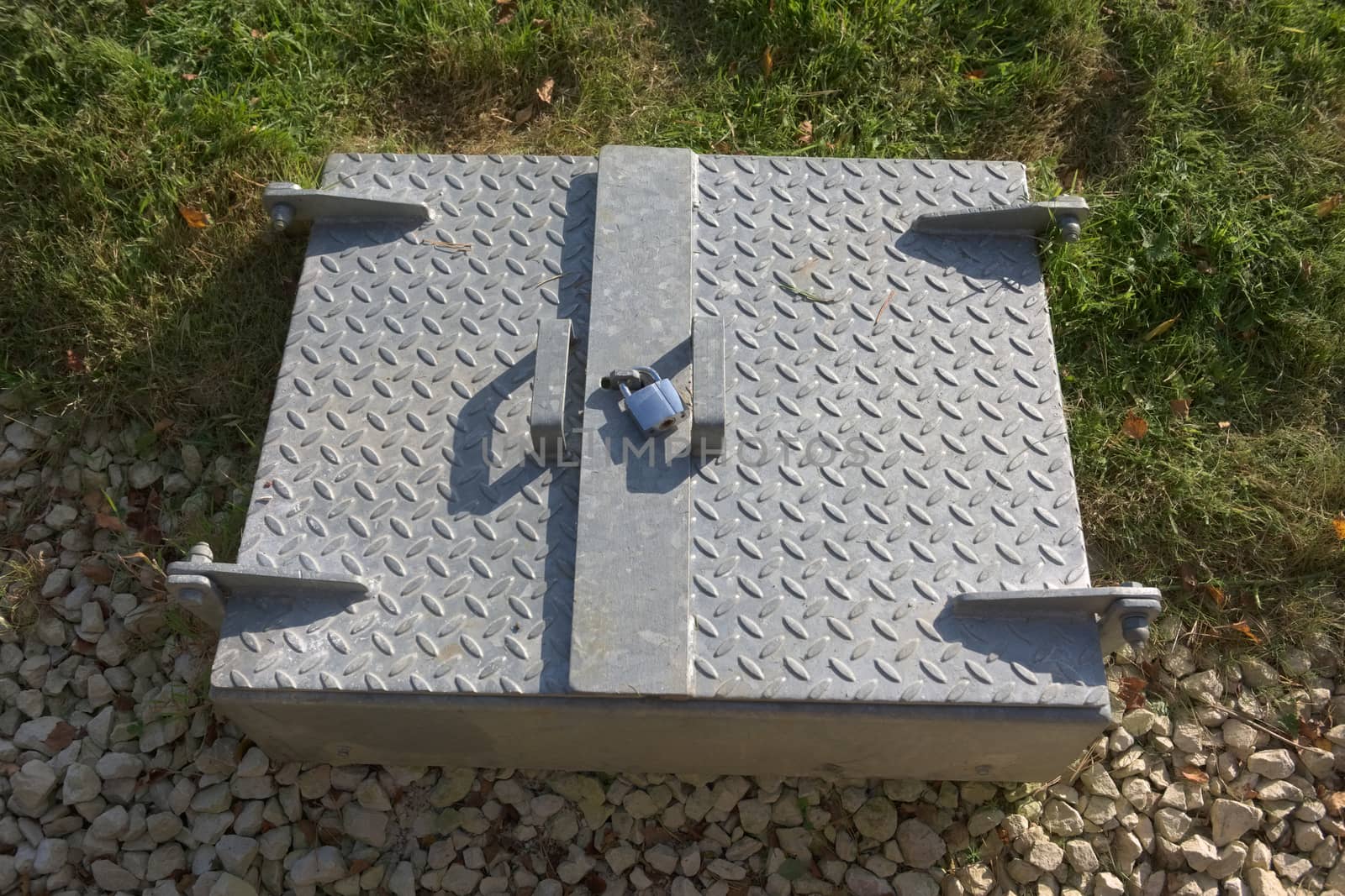 Modern secure padlocked steel service hatch in rural sunlight. Manhole cover juxtaposed against natural surroundings. Shadowed checkered steel texture