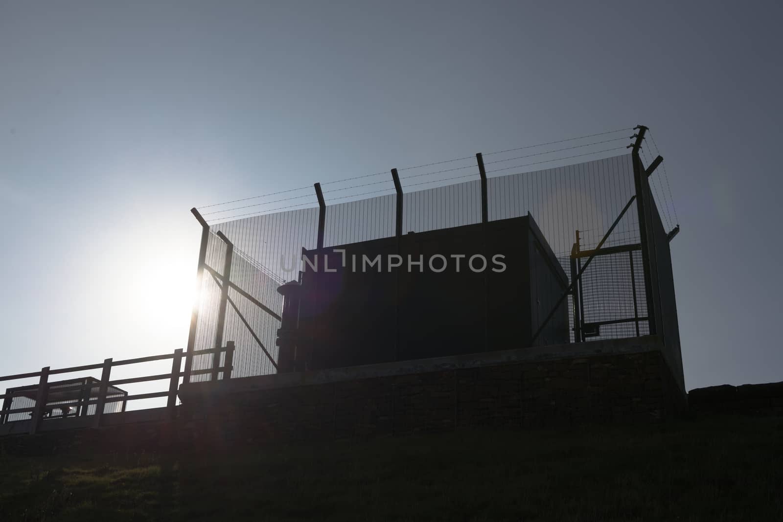 Tall barbed wire anti climb fence encases large square metal industrial water management equipment. Looking up, plain blue sky and bright sun behind