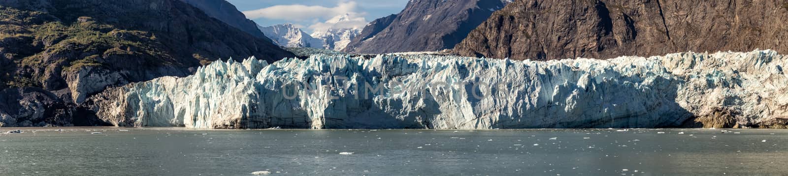 Panoramic view of Margerie glacier in Glacier Bay National Park and Preserve, Alaska, United States. Snowy mountain peaks and cloudy blue sky in the background.