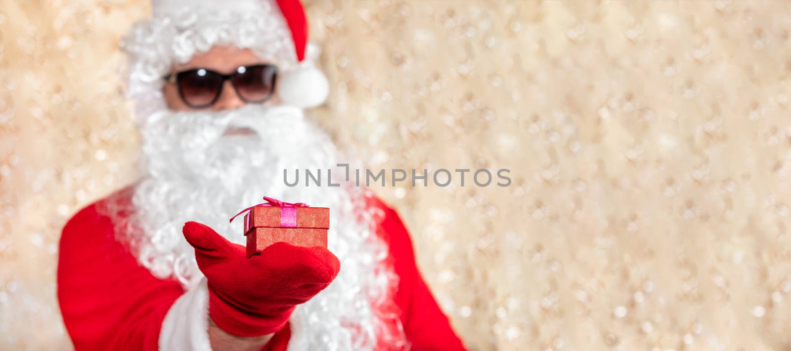 Santa Claus holding a Christmas present, red box with ribbon. Santa wearing sunglasses, with a long white beard, and is out of focus. Golden blurred sparkling background with copy space. Banner size by DamantisZ