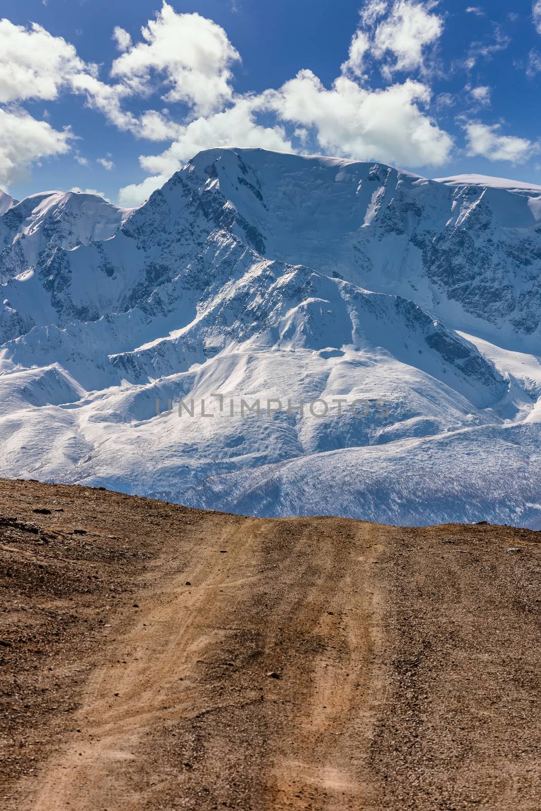 Portrait size landscape of a highland road leading to the snowy mountains of The North Chuyskiy mountain range. Beautiful blue cloudy sky as a background. Altai mountains, Siberia, Russia.