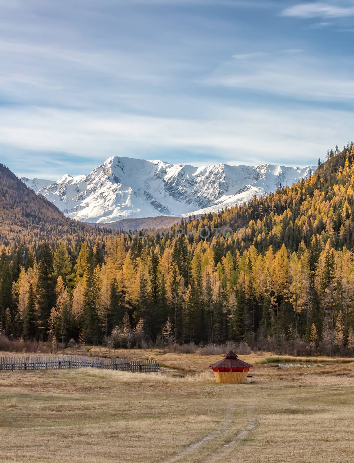 Scenic low angle view of snowy mountain peaks and slopes of North Chuyskiy ridge. Golden trees, wooden hut in the foreground. Beautiful blue cloudy sky as a backdrop. Altai mountains, Siberia, Russia by DamantisZ