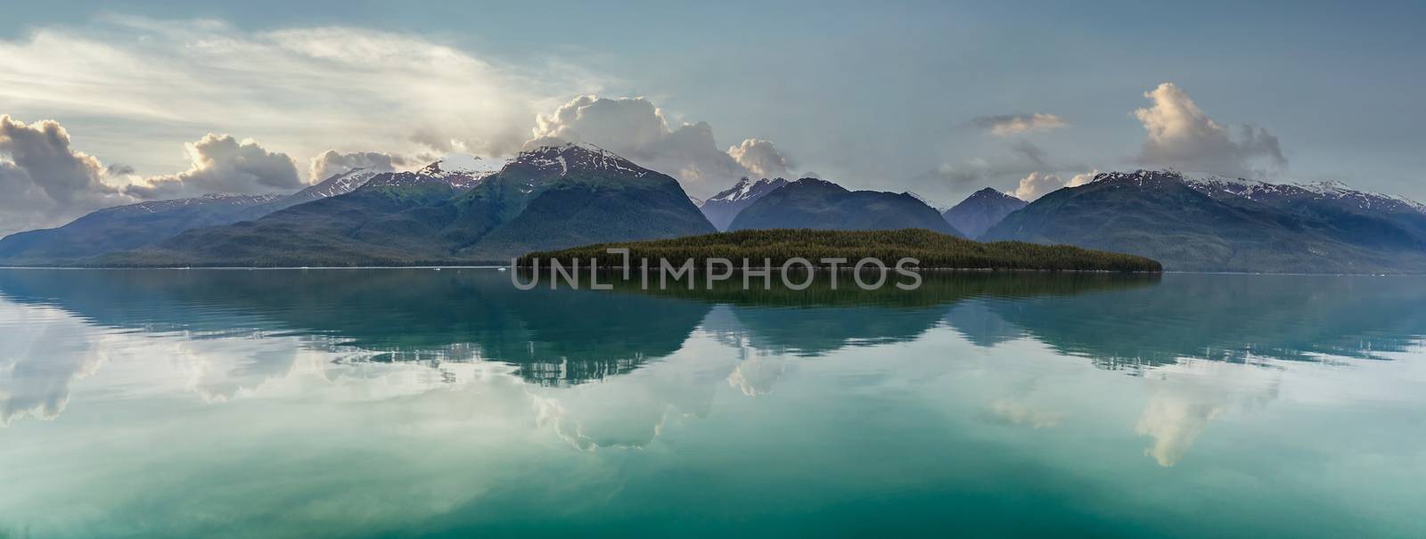 Beautiful landscape of snow capped mountains and a lone island full of green trees in a fjord in Alaska, USA. Cloudy sunset sky as a background. Mountains and sky reflecting in the water.