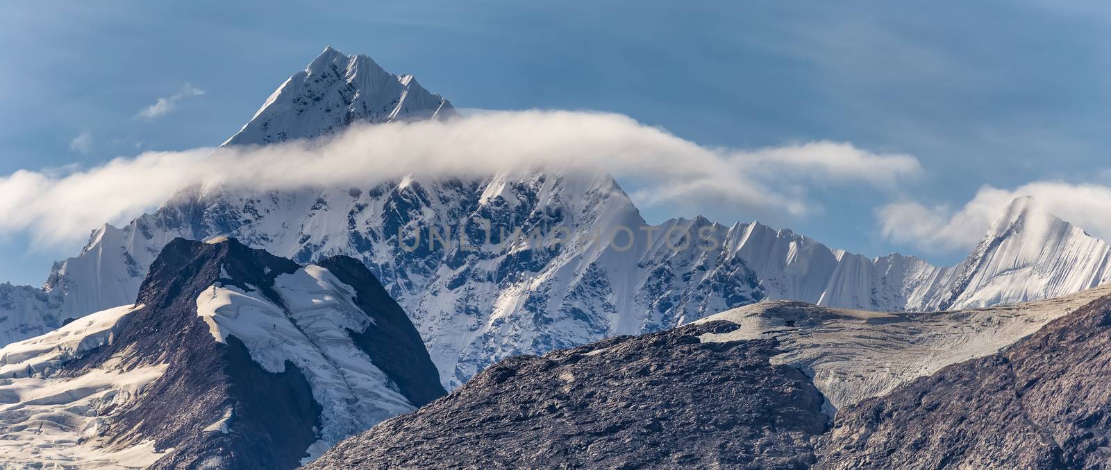 Beautiful panoramic view of a mountain peak covered with ice and snow, surrounded with fluffy couds in Alaska. Blue sky as a background by DamantisZ