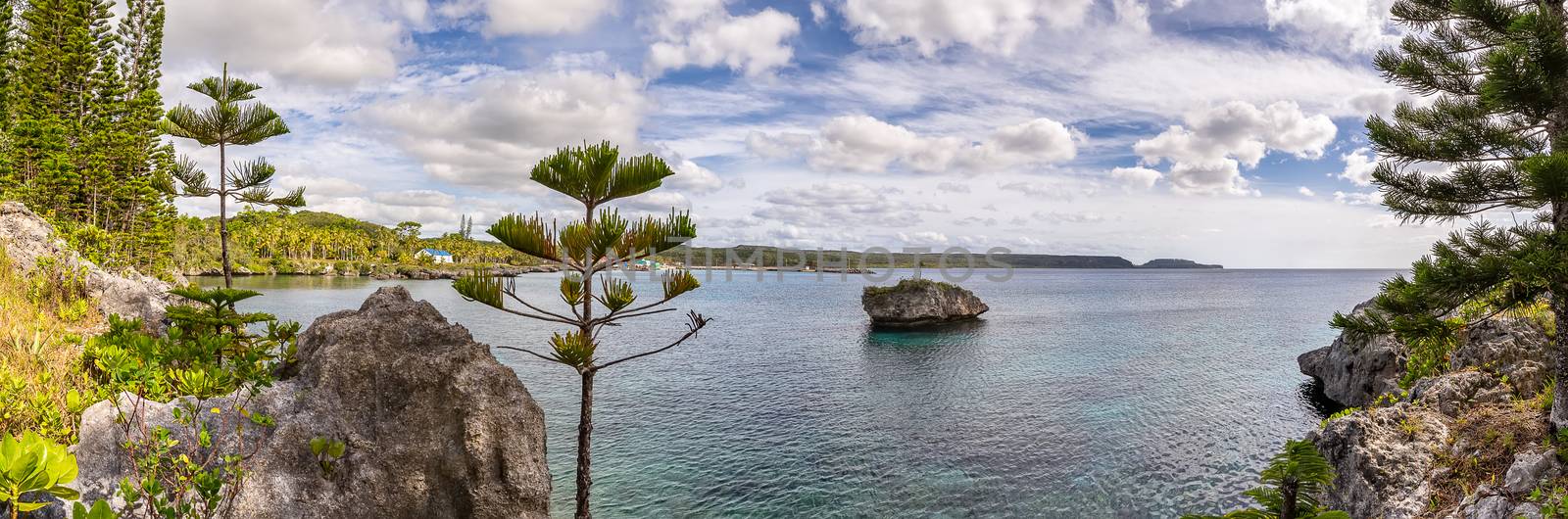 Amazing panoramic shot of Island of Mare in New Caledonia. Lone massive cliff in the water next to the island. Trees and rocks in the foreground. Blue cloudy sky as a background by DamantisZ