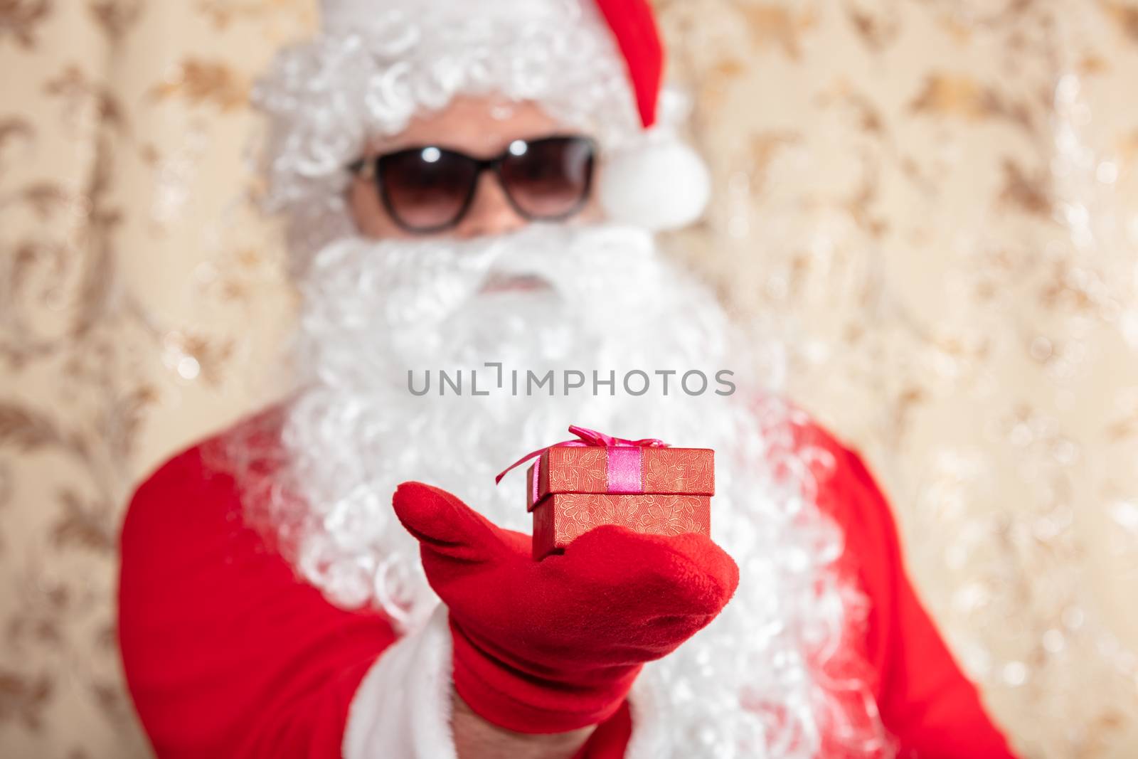Santa Claus holding a Christmas present, a red box with a ribbon. Santa wearing sunglasses, with a long white beard, and is out of focus. Golden blurred sparkling background.