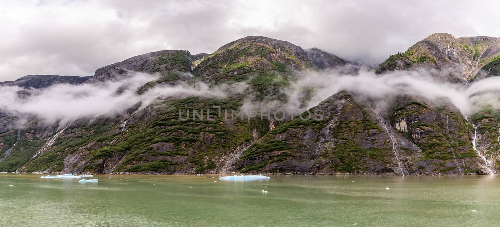 Beautiful panoramic view of mountains covered with fluffy clouds and trees in Alaskan fjord. Cloudy sky as a background.