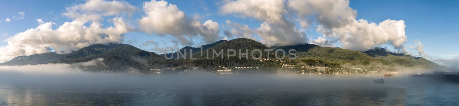 Panoramic shot of mountains covered with clouds and fog in Gastineau Channel, Juneau, Alaska. Golden hour. Blue cloudy sky as a background and boat sailing in the fog in the foreground.