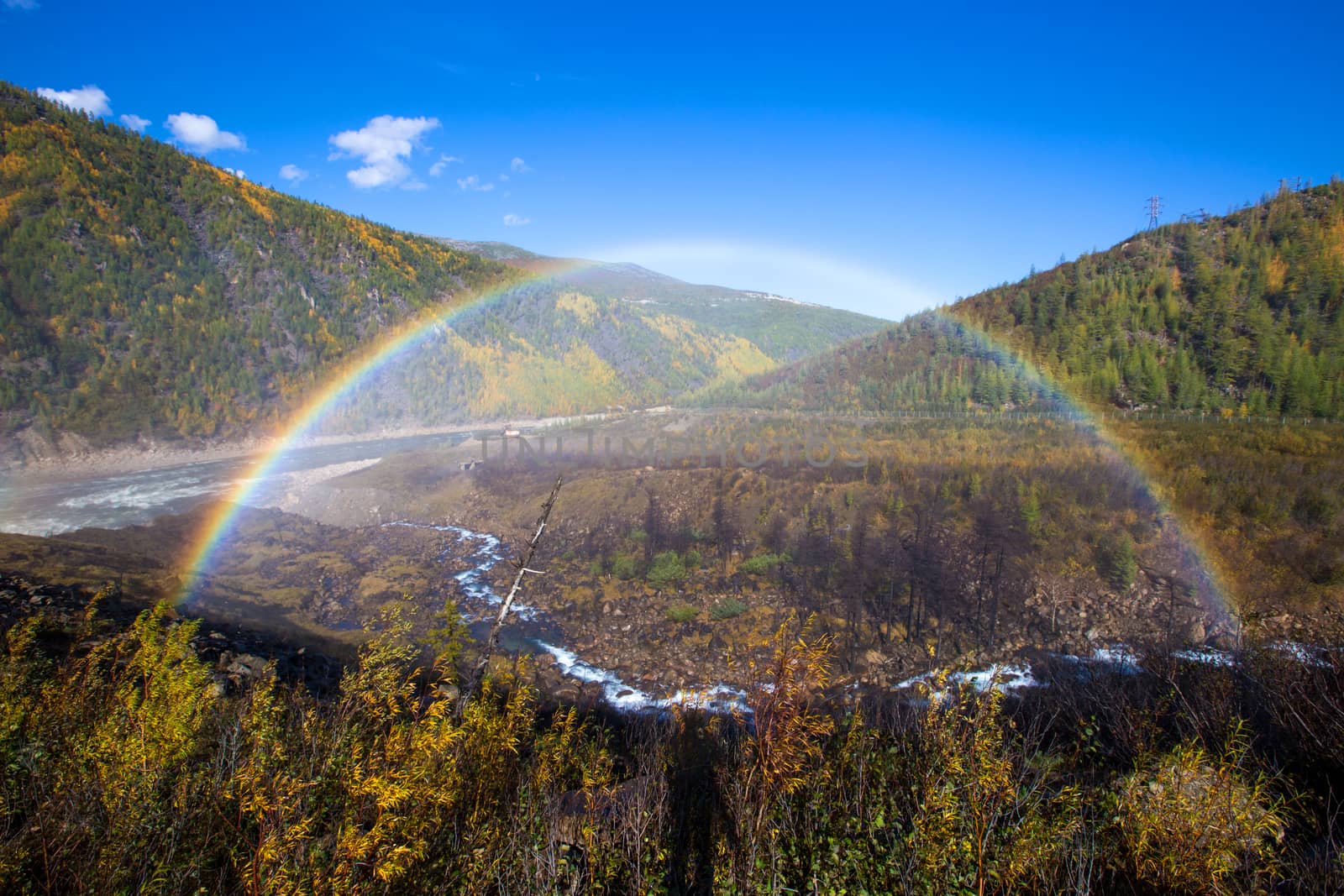 A beautiful bright rainbow in full size against the background of autumn hills and a small stream.