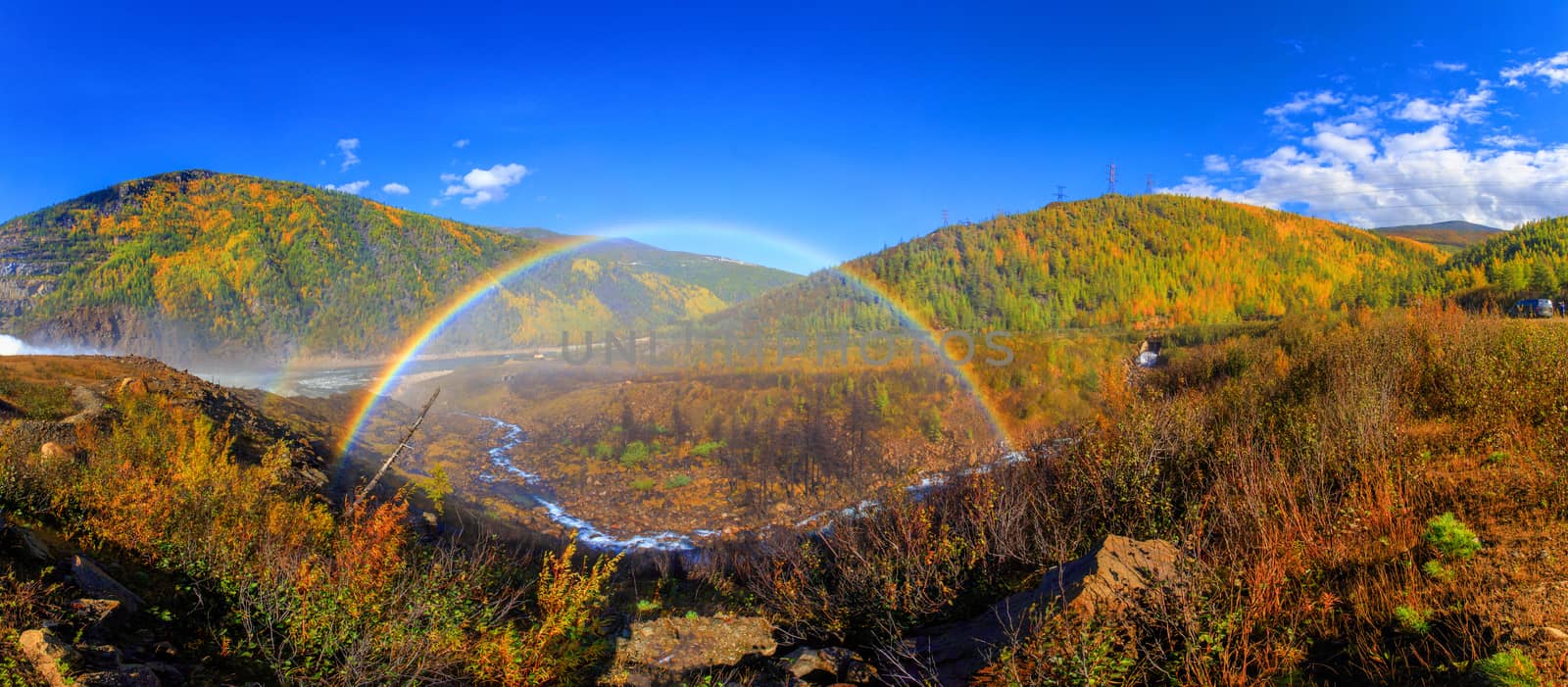 A beautiful bright rainbow in full size against the background of autumn hills and a small stream. Rainbow on hydroelectricity during spillway by PrimDiscovery