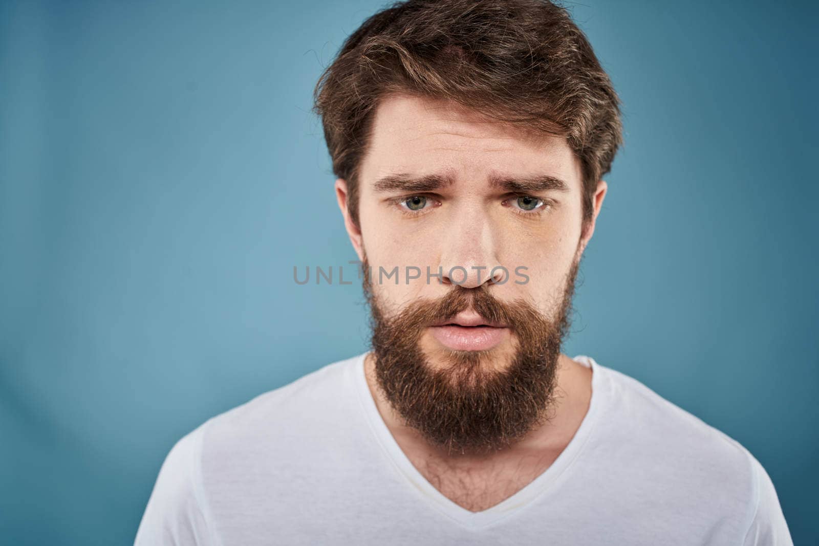 Bearded man displeased facial expression emotions close-up blue background white t-shirt by SHOTPRIME