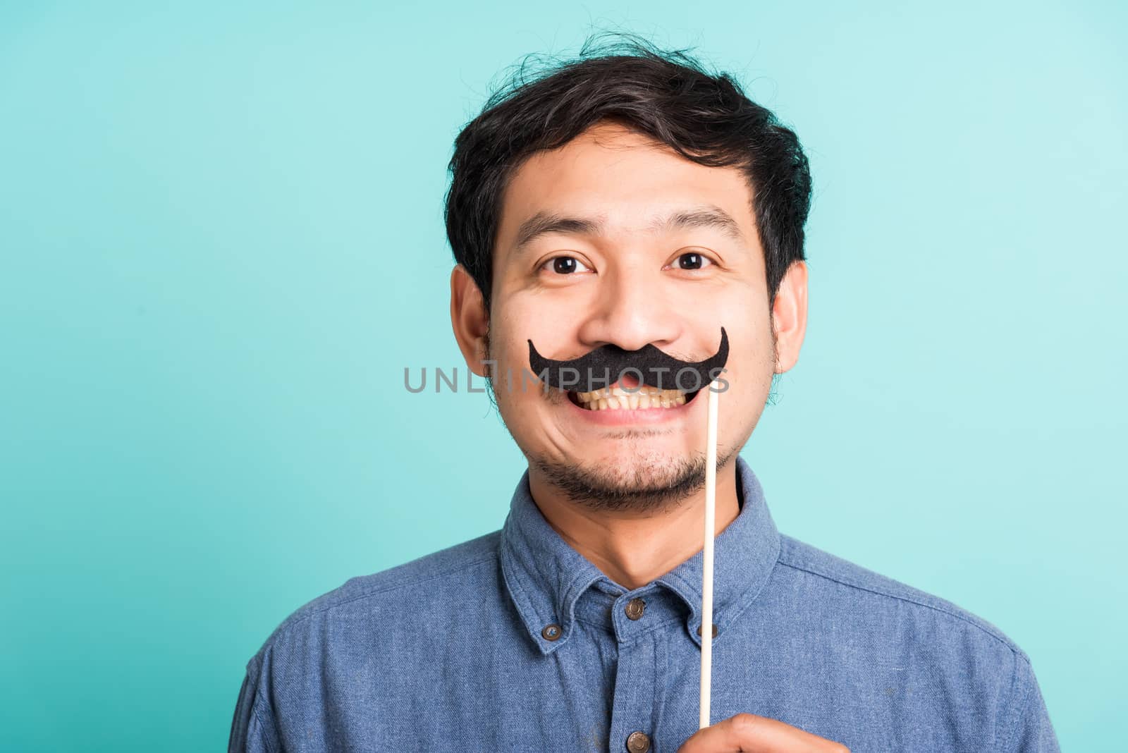 Portrait Asian happy handsome man posing he holding a funny mustache card or vintage fake moustaches on his mouth, studio shot isolated on blue background, Fathers day, November day concept