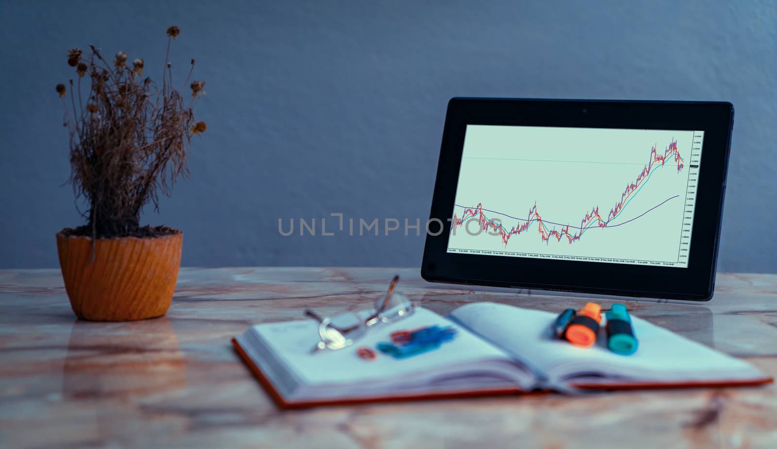 Screen of a tablet on a desk with a chart of currency pairs. On the desk there is an out of focus notebook