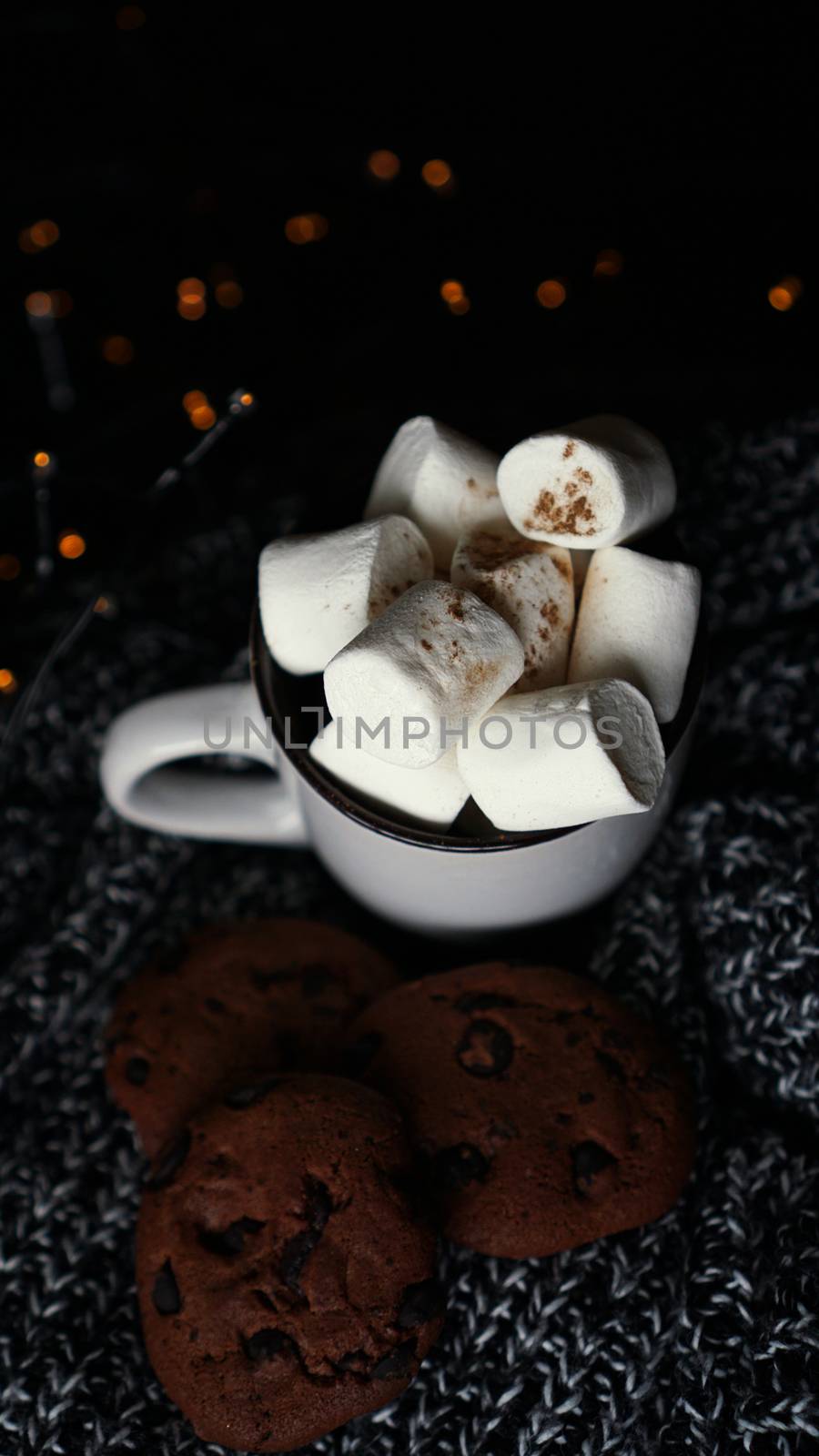 Mug with marshmallows and chocolate chip cookies on dark Christmas background with lights - vertical photo