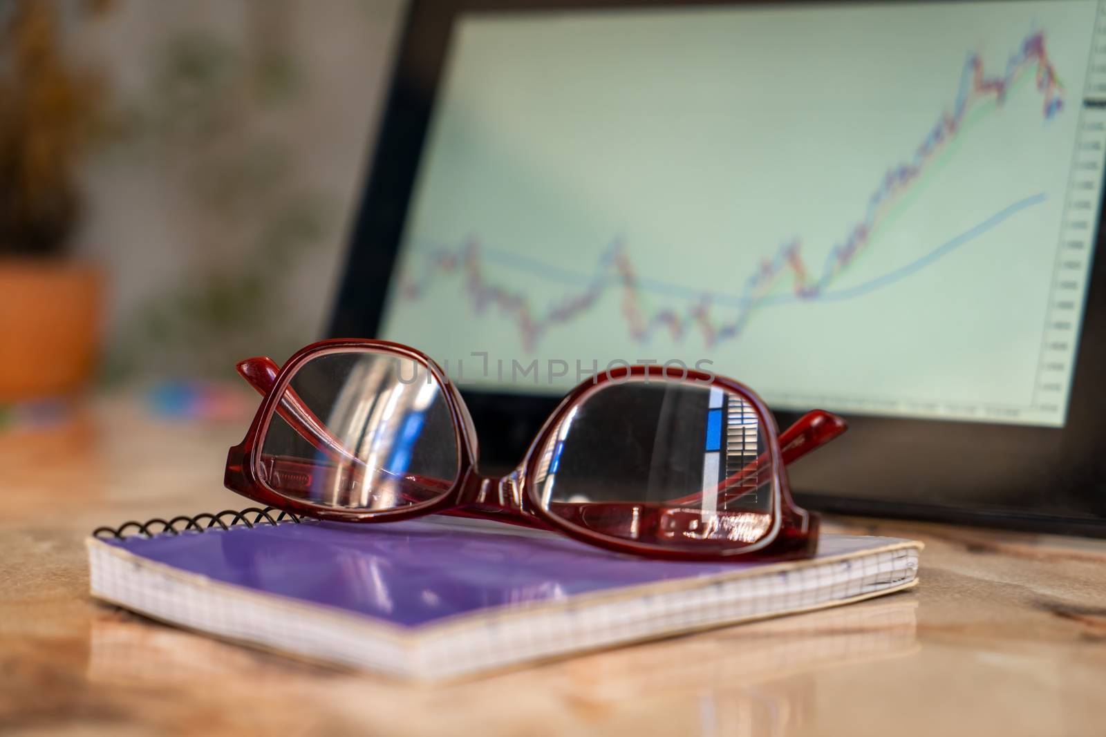 Close-up photo of an glasses on a notebook. There is a tablet screen out of focus with a graphic, in the background