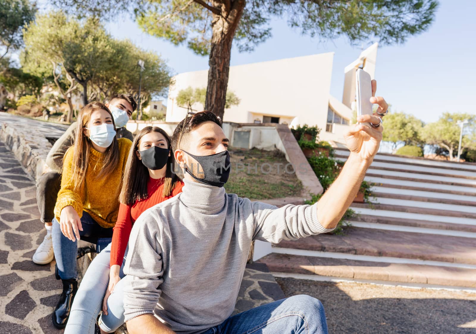 Group of young caucasian friends sitting on a city square wall wearing covid-19 coronavirus face masks making self portrait with smartphone for sharing on social networks - Focus on first man