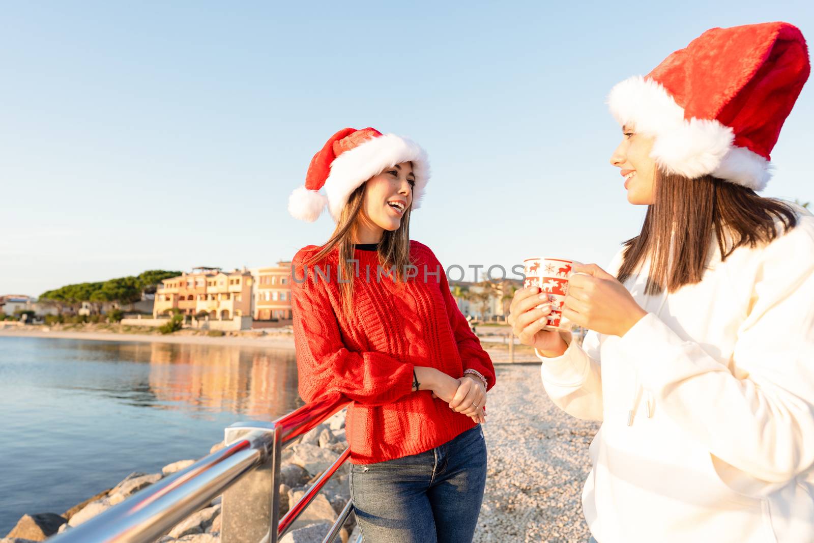 Beautiful young caucasian women standing outdoor talking with a Christmas mug in hands wearing Santa hat - Sunset sea winter vacation for two female friends at Golfo Aranci in Costa Smeralda, Sardinia by robbyfontanesi