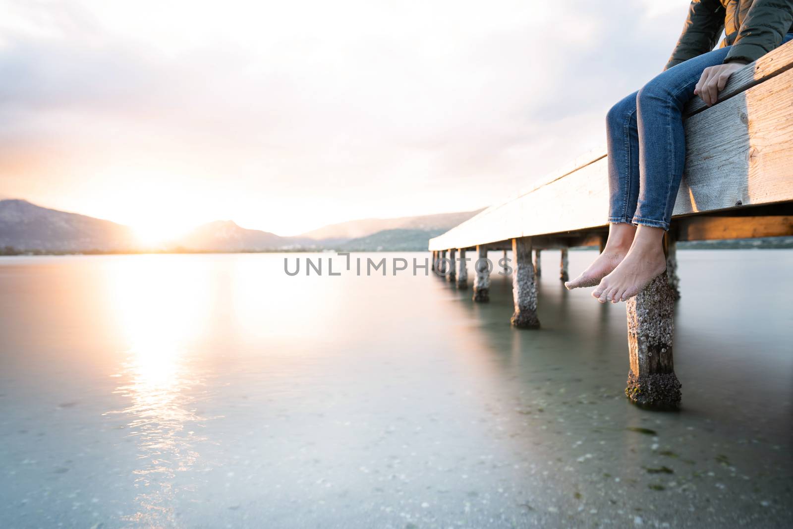 Alone unrecognizable female person sitting on a wooden pier at sunset or dawn with feet dangling on the water - Pensive woman with winter dress admires the sun going down in the evening on the water