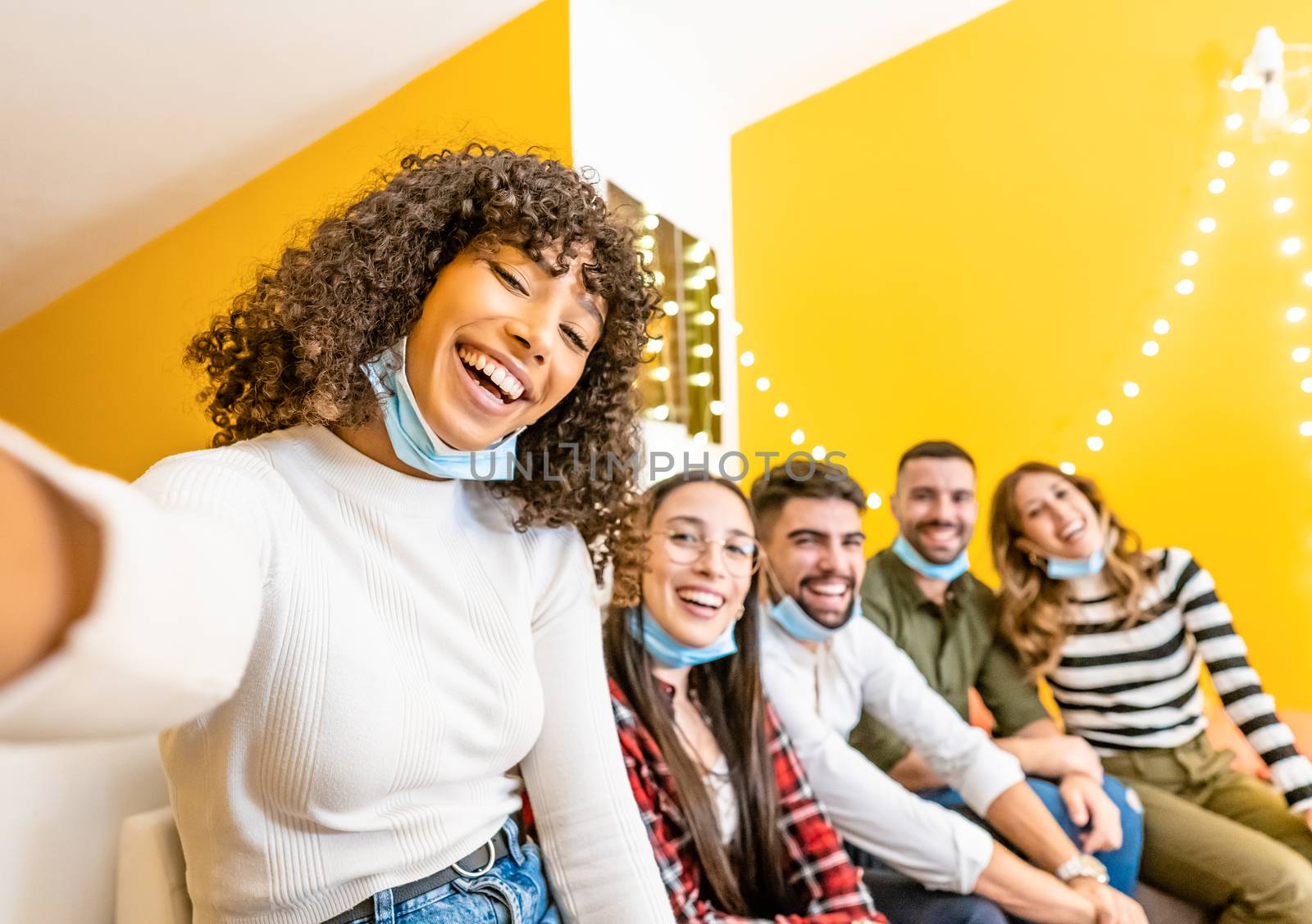 Group of multi ethnic young people having tech fun home - Beautiful black Hispanic woman take a photo with smartphone to her friends wearing lowered Coronavirus face mask protection - Focus on curly