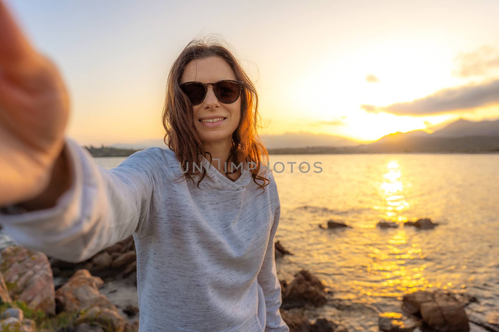 Young beautiful caucasian woman make a self portrait holding the camera at sunset on sea rocks - Smartphone selfie with water reflection of the setting sun on the water - Female alone vacation concept