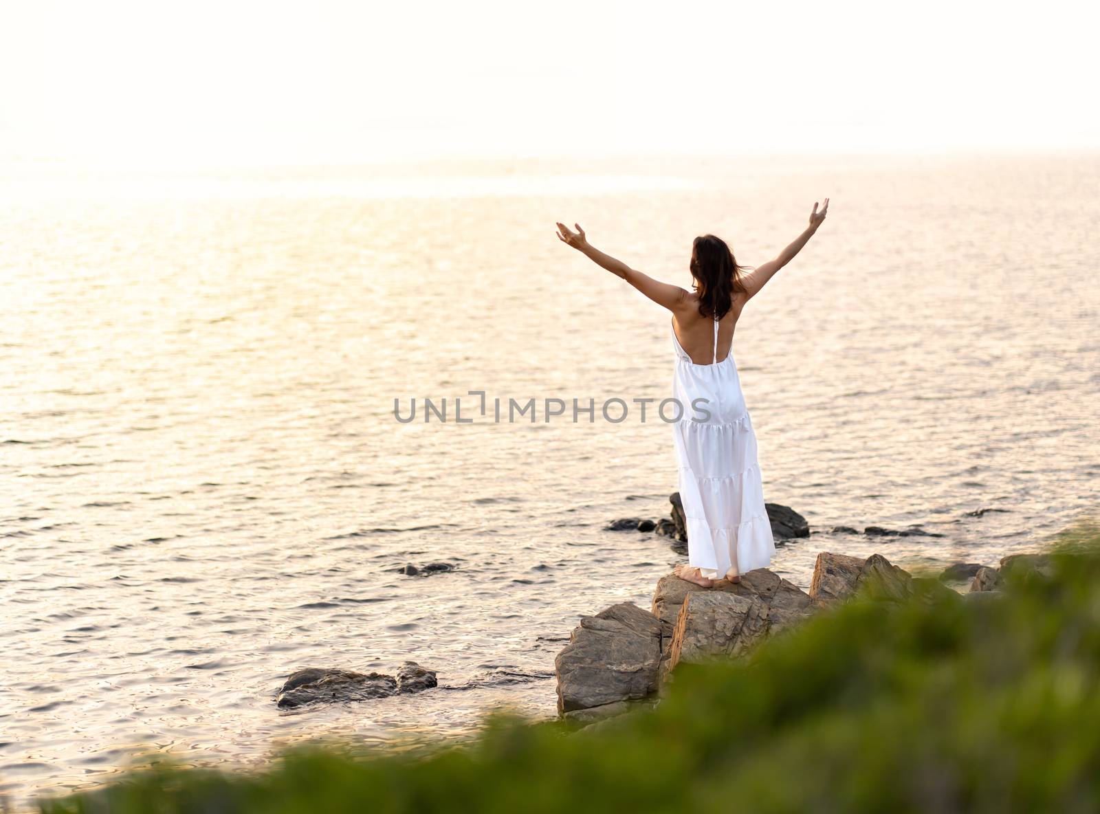 View from back of an unrecognizable woman with white long dress and open arms upwards looking at the setting sun - Alone female person with barefoot on sea rocks embracing nature as a sign of devotion