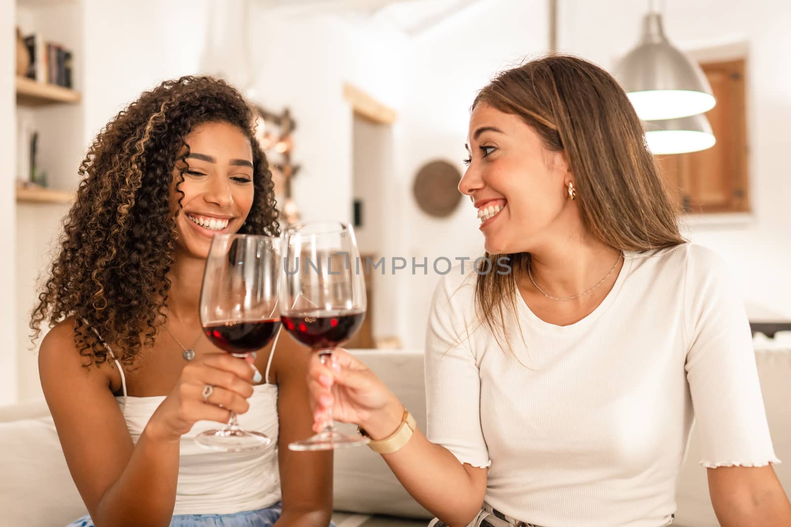 Two young mixed race woman celebrating at home toasting with red wine glasses - Smiling Hispanic curly brunette clinking with her best friend sitting at he sofa in her modern elegant living room