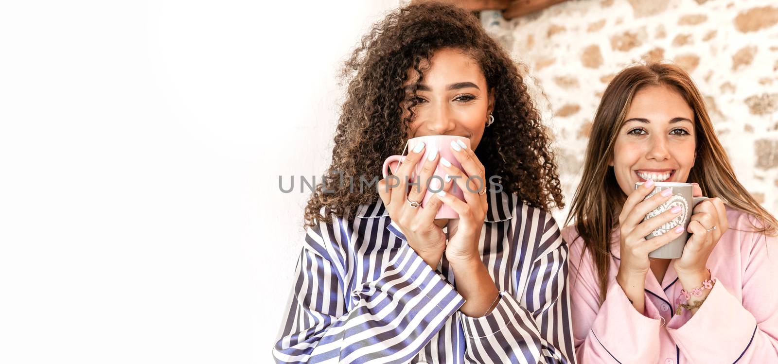 Mixed race female couple in pajama just woken up standing outdoor drinking tea looking at the camera - Two women friends smiling holding a mug with large white copy space to the left by robbyfontanesi