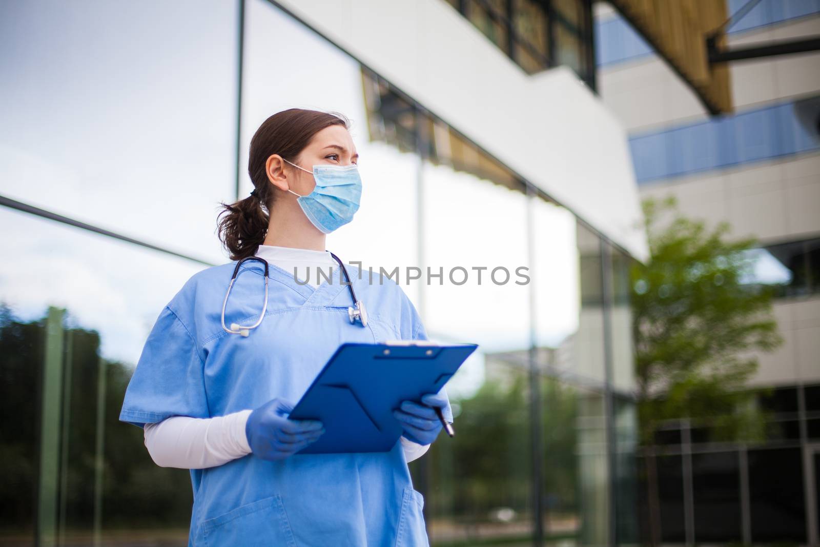 Young female caucasian UK NHS doctor looking in distance with fear worry anxiety & uncertainty in eyes,wearing uniform & face mask,hope & faith & overcoming global Coronavirus COVID-19 pandemic crisis