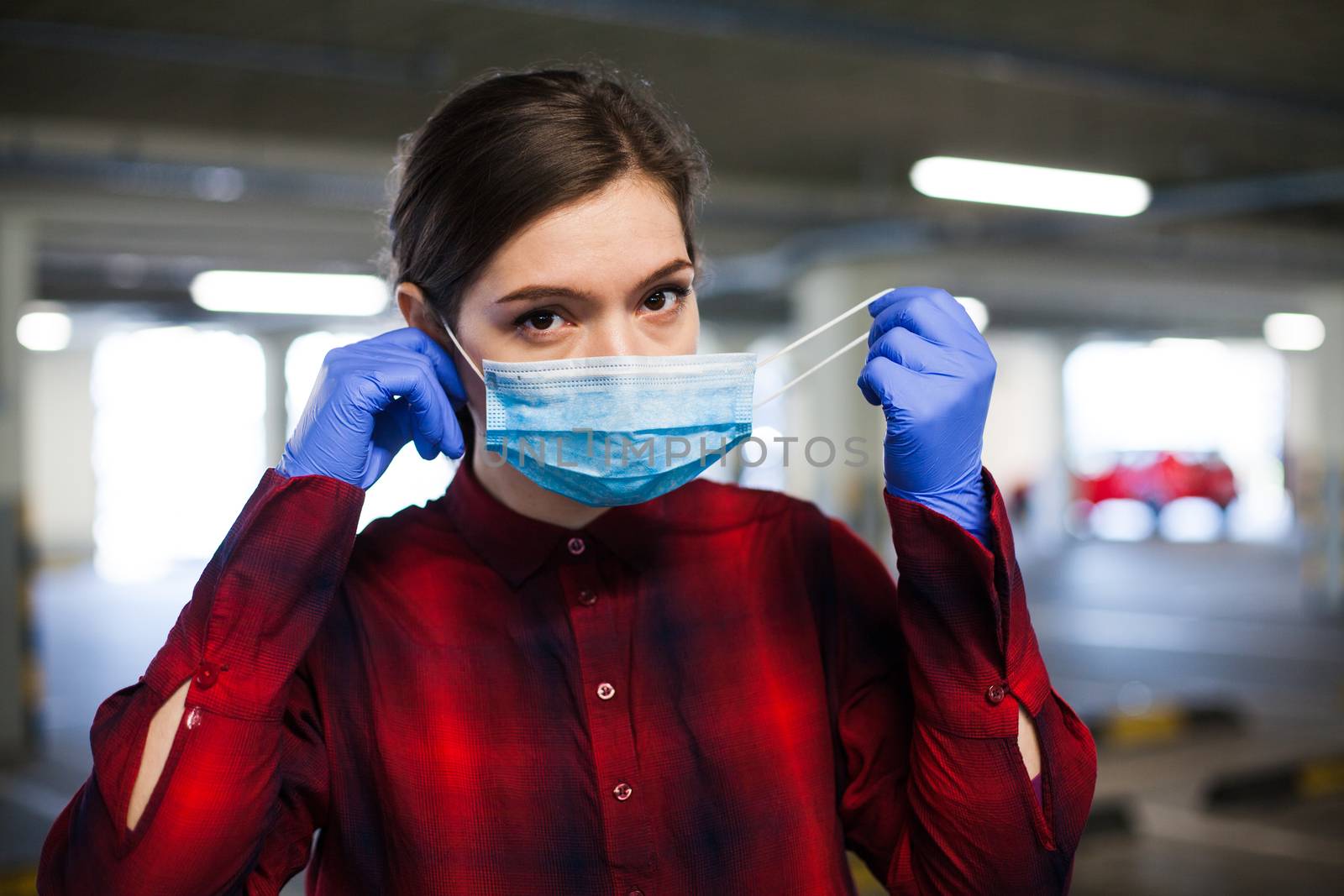 Young caucasian woman wearing surgical gloves putting face mask on, protection from spread of Coronavirus COVID-19 corona virus disease infection, portrait in empty underground garage car parking lot 