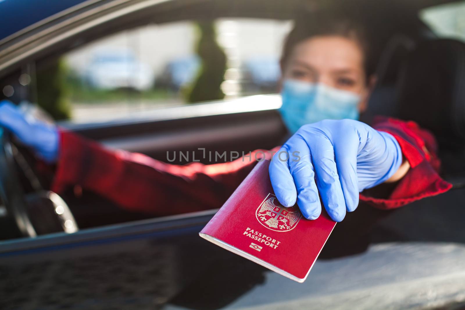 Young caucasian female driver wearing medical face mask and protective gloves handing over red immunity passport through car window,safety security border health check,Coronavirus COVID-19 pandemic