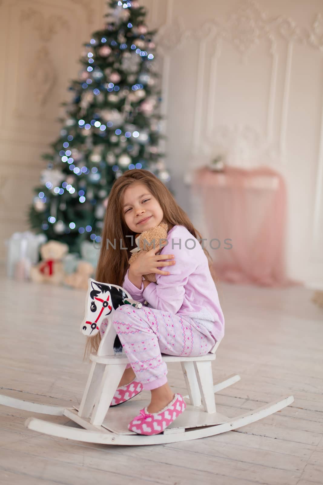 A beautiful little girl in pink pajamas rejoices in a wooden rocking horse, a gift from Santa for Christmas.