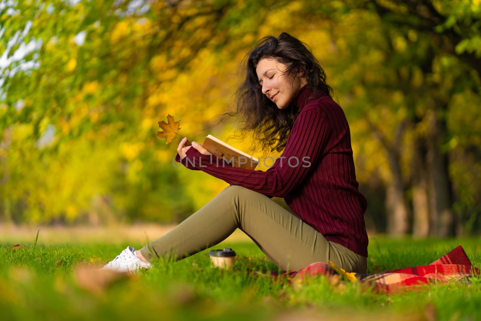 A cute girl reads a book and drinks coffee on a green lawn in an autumn park. Autumn mood. A cozy place to be alone with yourself.