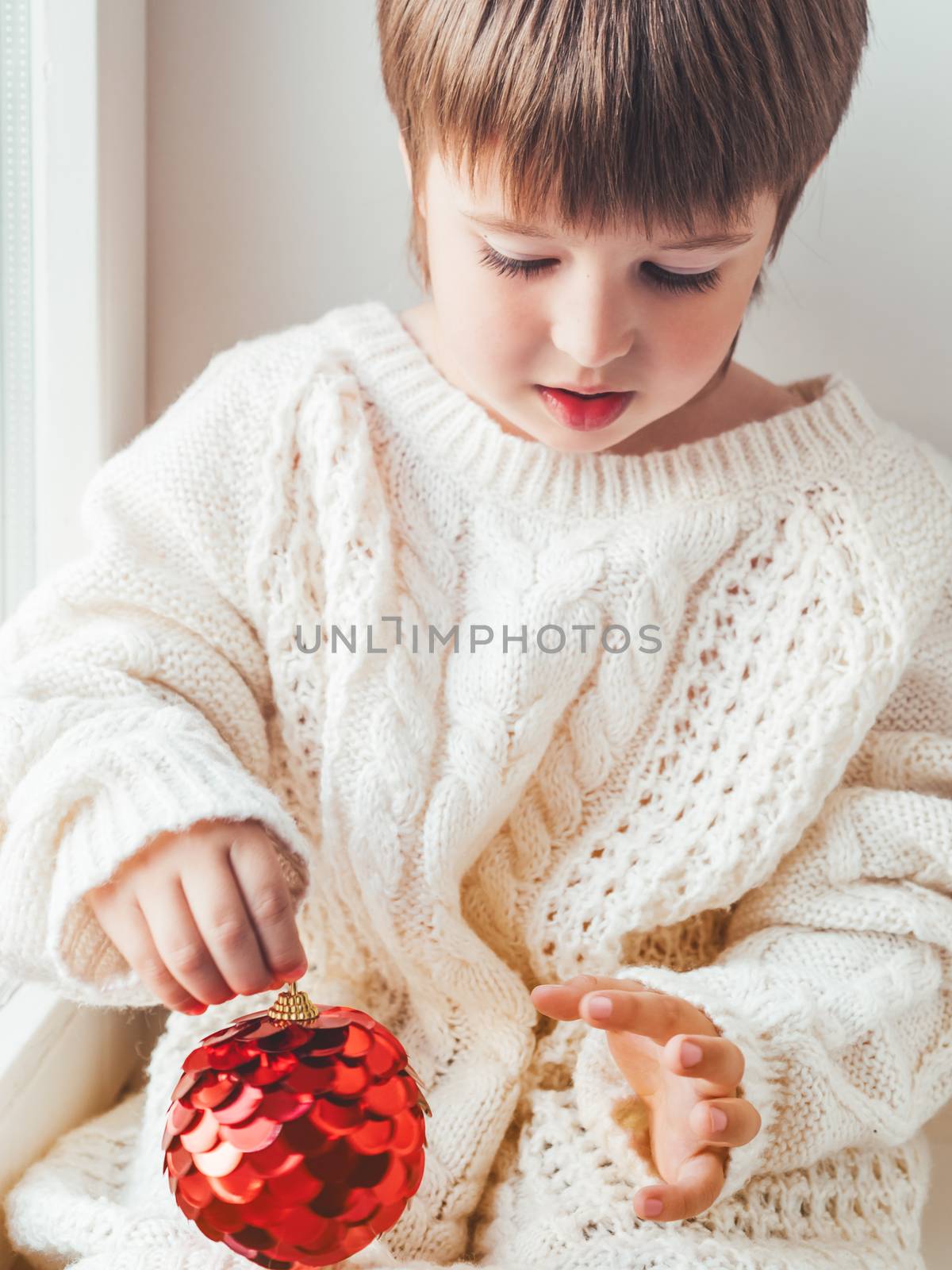 Kid with red decorative ball for Christmas tree. Boy in cable-kn by aksenovko
