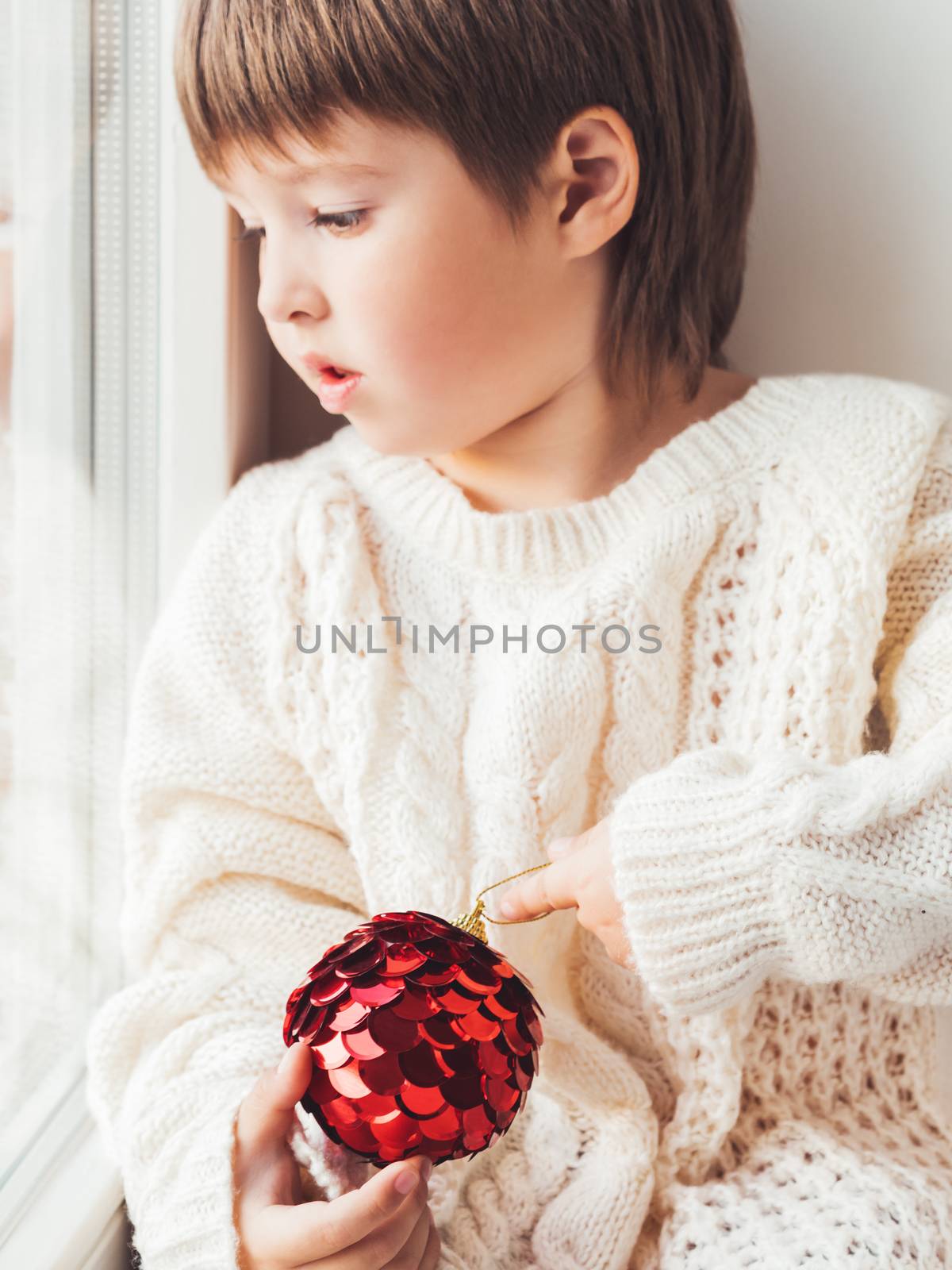 Kid with red decorative ball for Christmas tree. Boy in cable-knit oversized sweater. Cozy outfit for snuggle weather. Winter holiday spirit. New year.