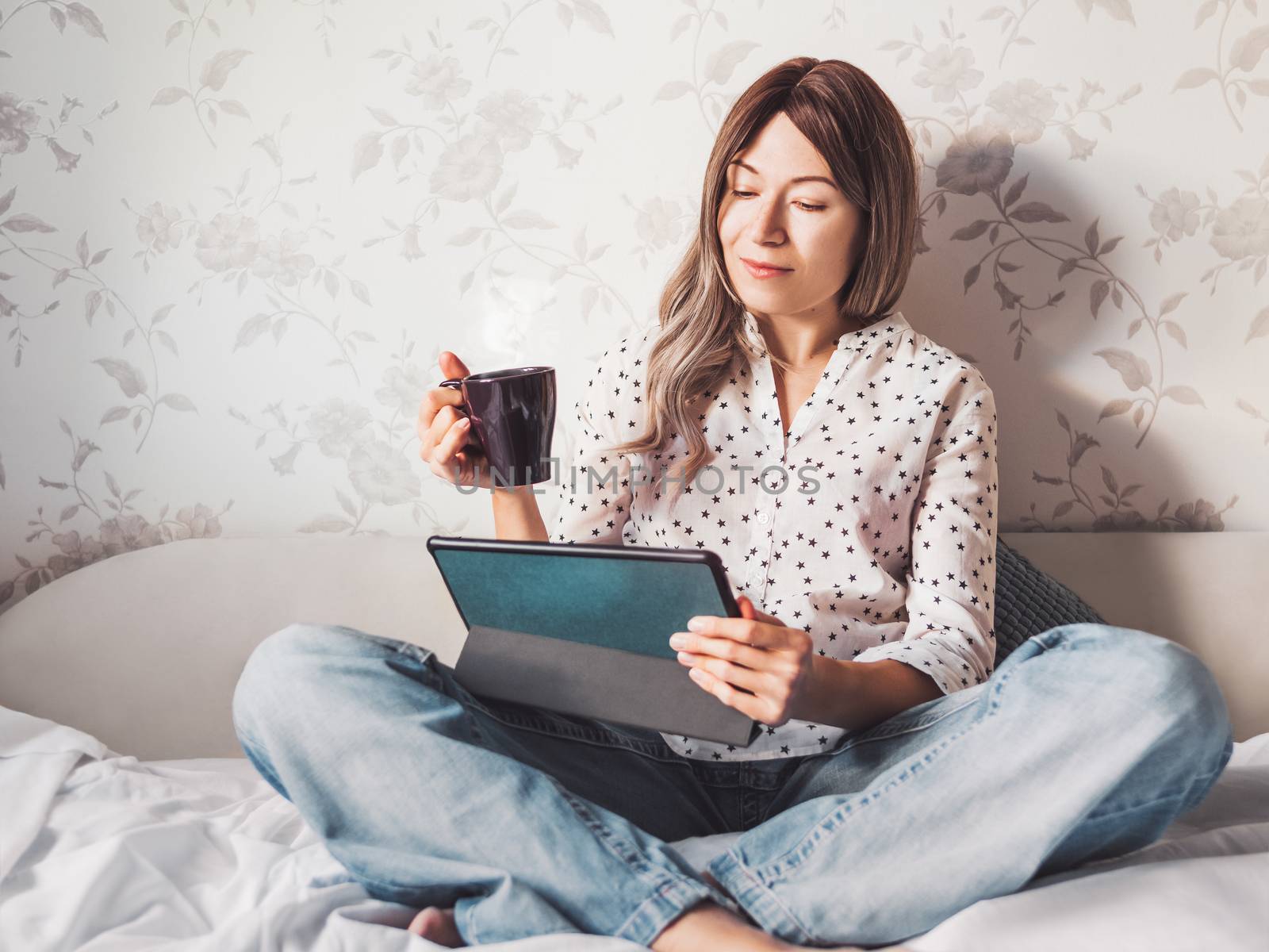 Woman sits on bed with tablet PC and cup of hot coffee. She watches online TV series. Online video call or conference, distance learning, remote education. Self isolation during quarantine.