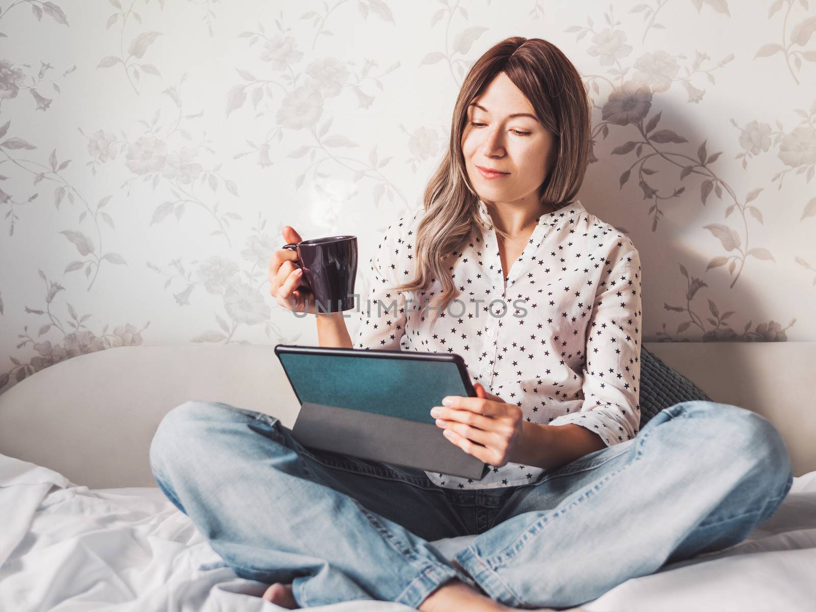 Woman sits on bed with tablet PC and cup of hot coffee. She watc by aksenovko