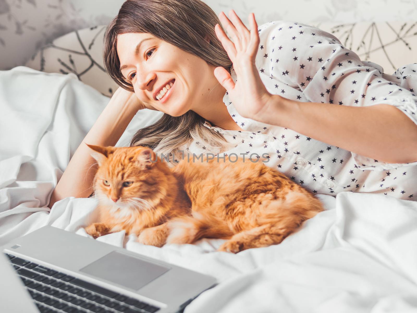 Cute ginger cat and woman are lying in bed. Woman is greeting somebody via online video translation or video call. Online communication. Morning bedtime with fluffy pet.