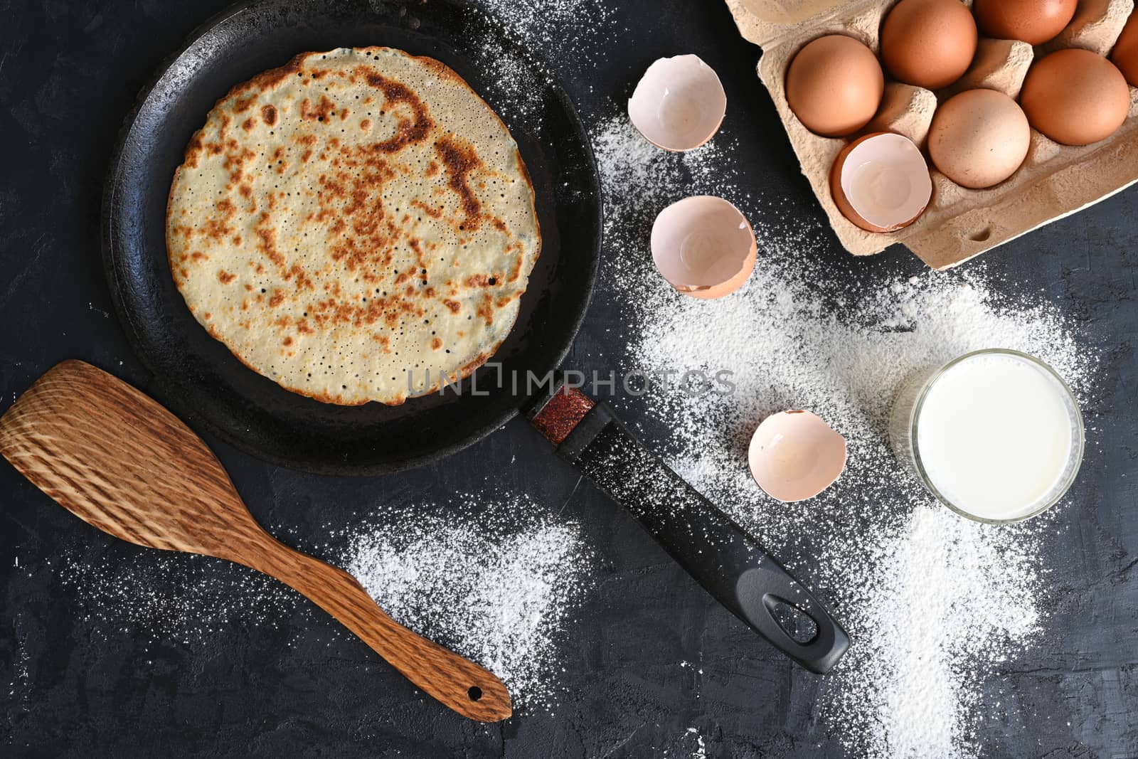 Hot pancake in black pan on black table with flour, milk and eggs.