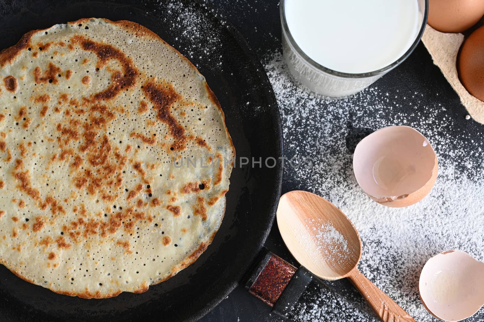 Hot pancake in black pan on black table with flour, milk and eggs.