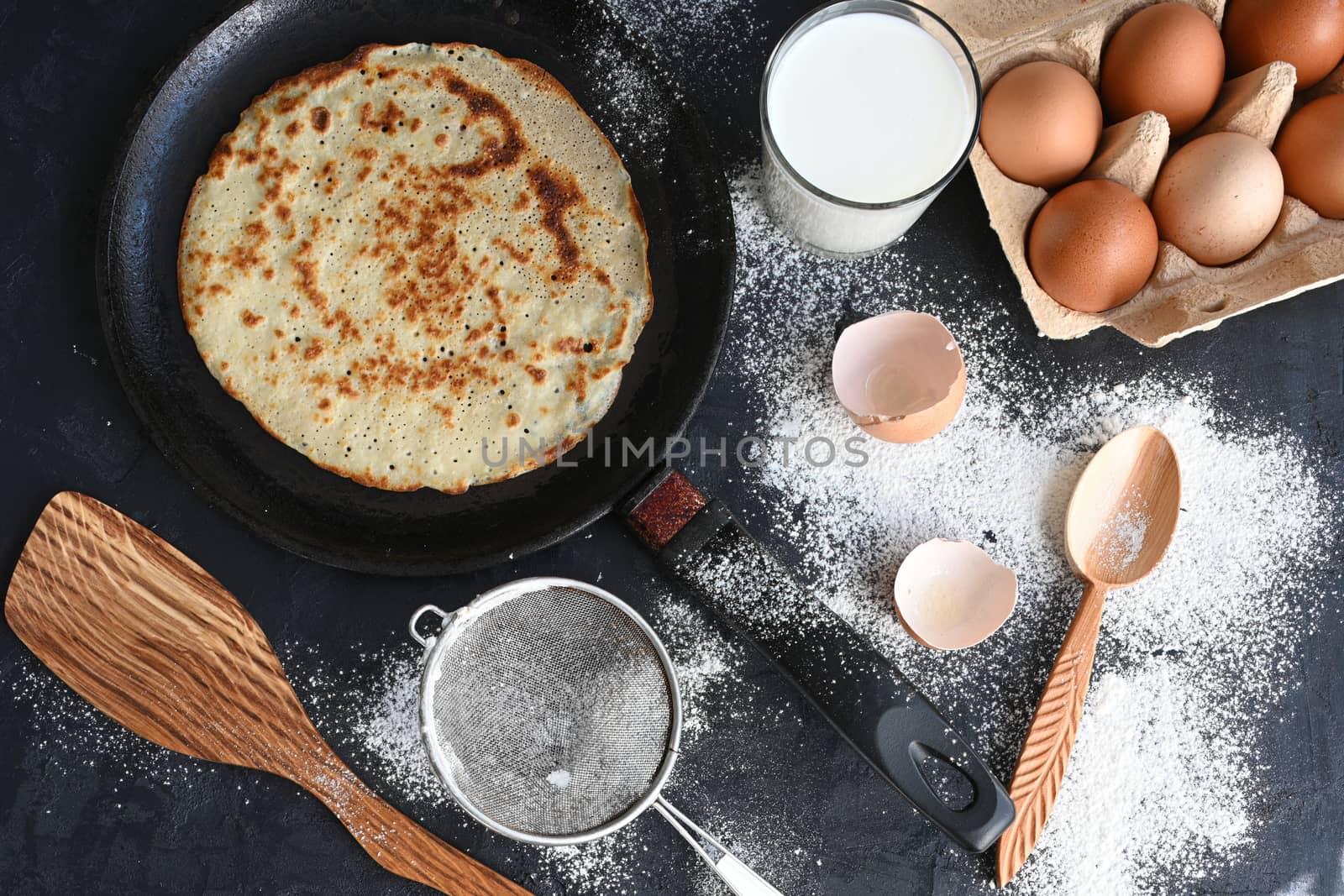 Hot pancake in black pan on black table with flour, milk and eggs.