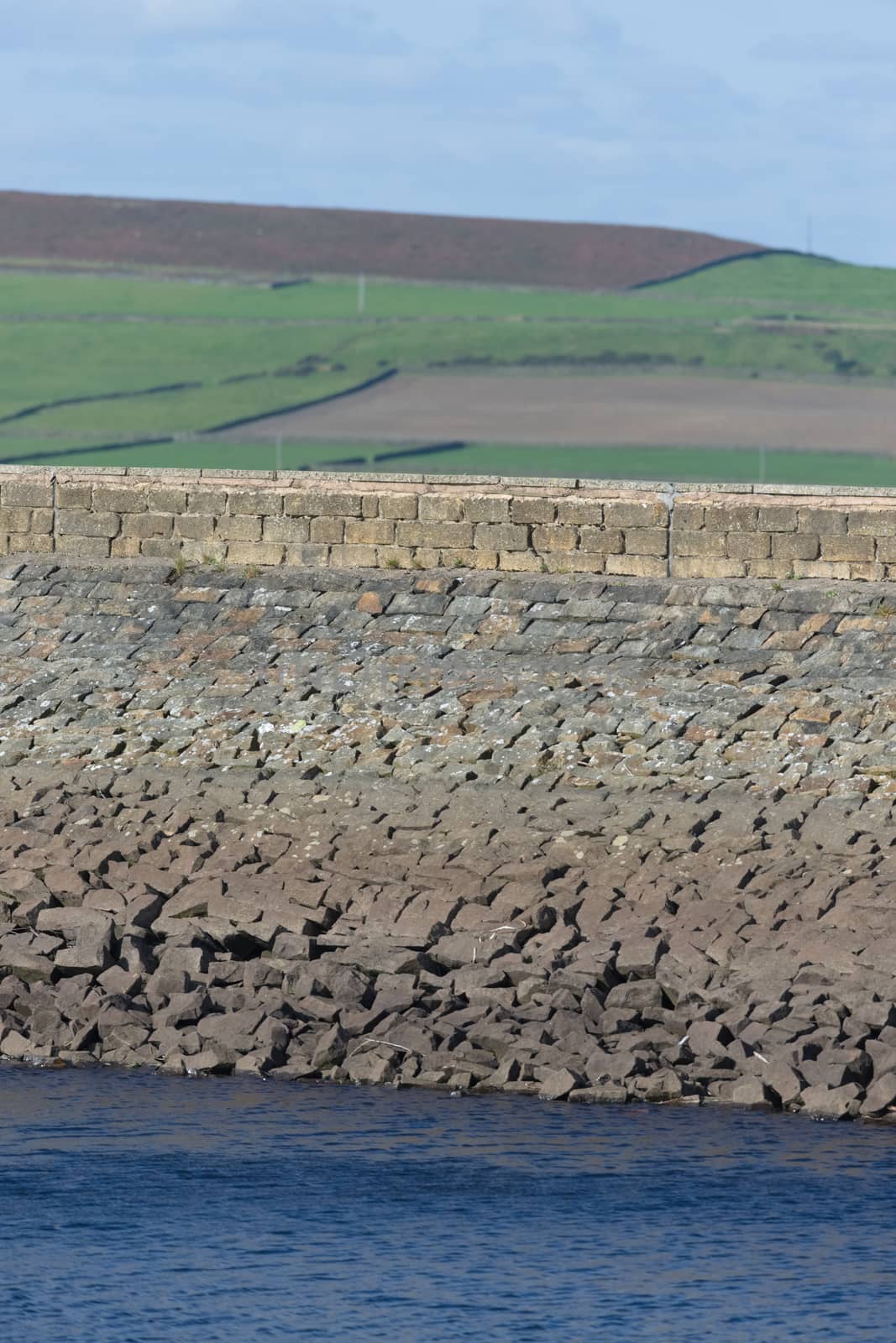 Vertical image of stone dam reservoir wall. Deep blue water and rural agricultural background are selectively out of focus. Warm sunny summers day