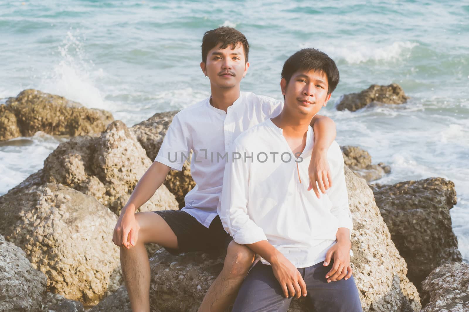 Homosexual portrait young asian couple sitting hug together on rock or stone in the beach in summer, asia gay going tourism for leisure and relax with happy in vacation at sea, LGBT legal concept.