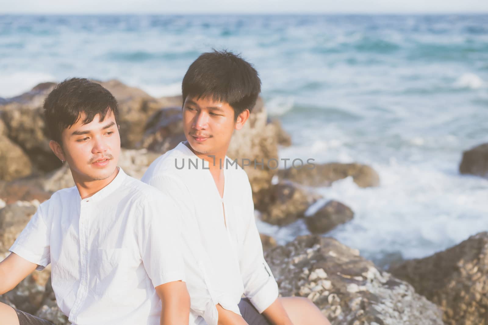 Homosexual portrait young asian couple sitting hug together on rock or stone in the beach in summer, asia gay going tourism for leisure and relax with happy in vacation at sea, LGBT legal concept.