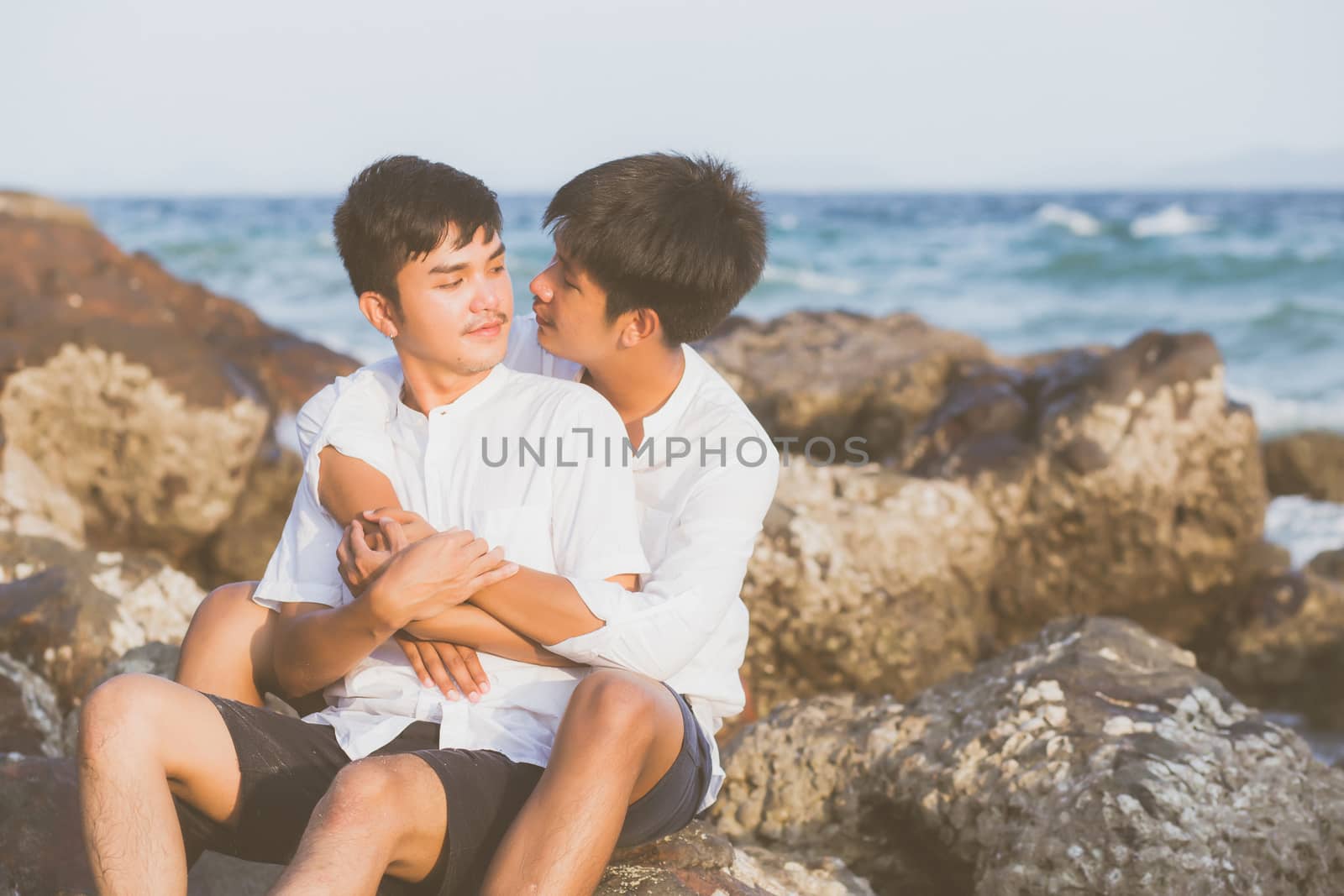 Homosexual portrait young asian couple sitting hug together on rock or stone in the beach in summer, asia gay going tourism for leisure and relax with happy in vacation at sea, LGBT legal concept.