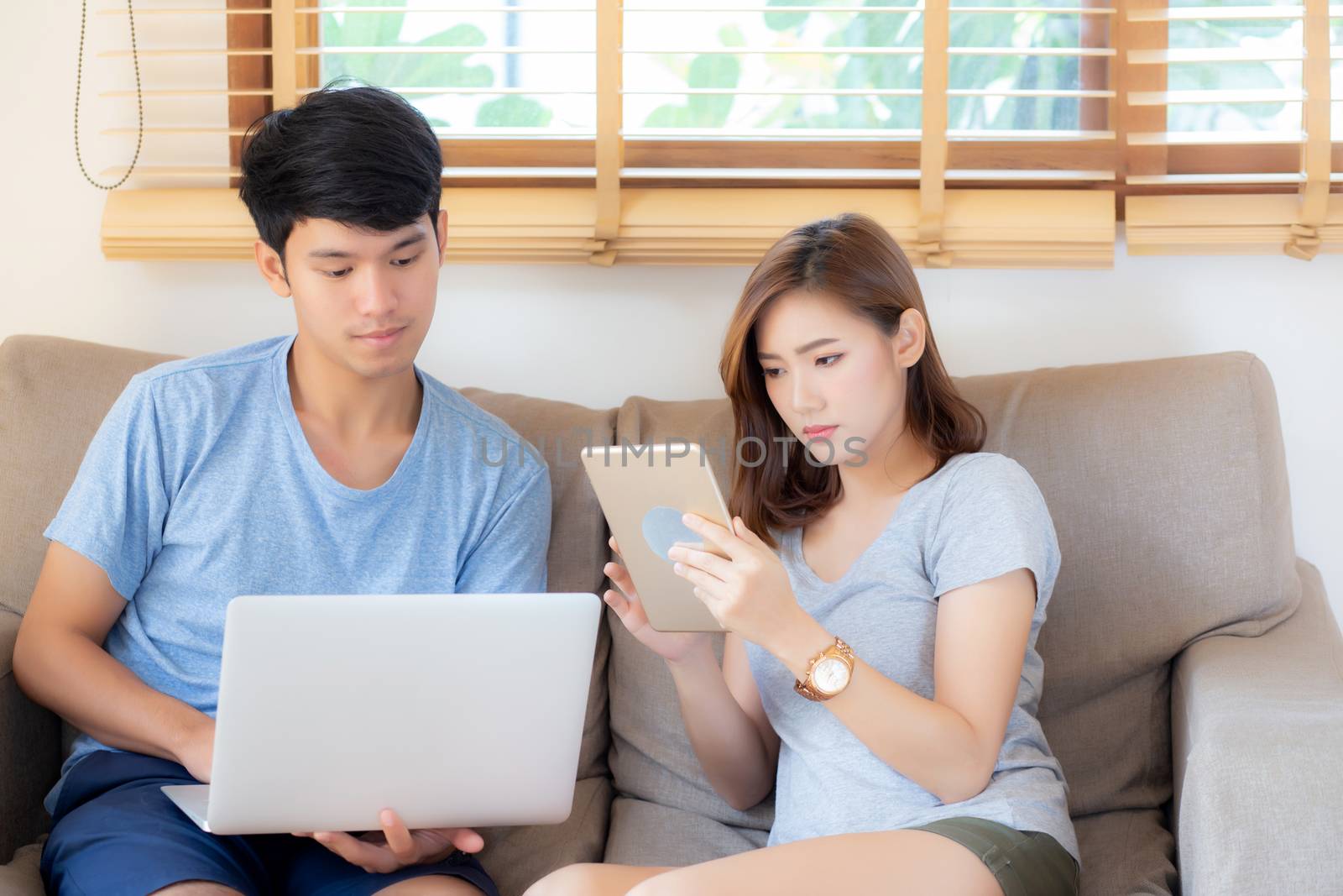 Beautiful young asian couple cheerful freelance working with man using laptop and woman using tablet on couch, family relax and sharing communication together at home, lifestyle concept.